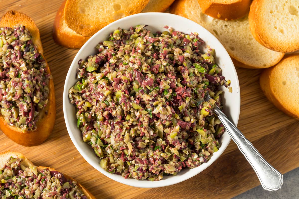 A bowl of chopped olive salad with green and black olives, giardiniera, roasted red peppers, capers, and herbs on a kitchen counter