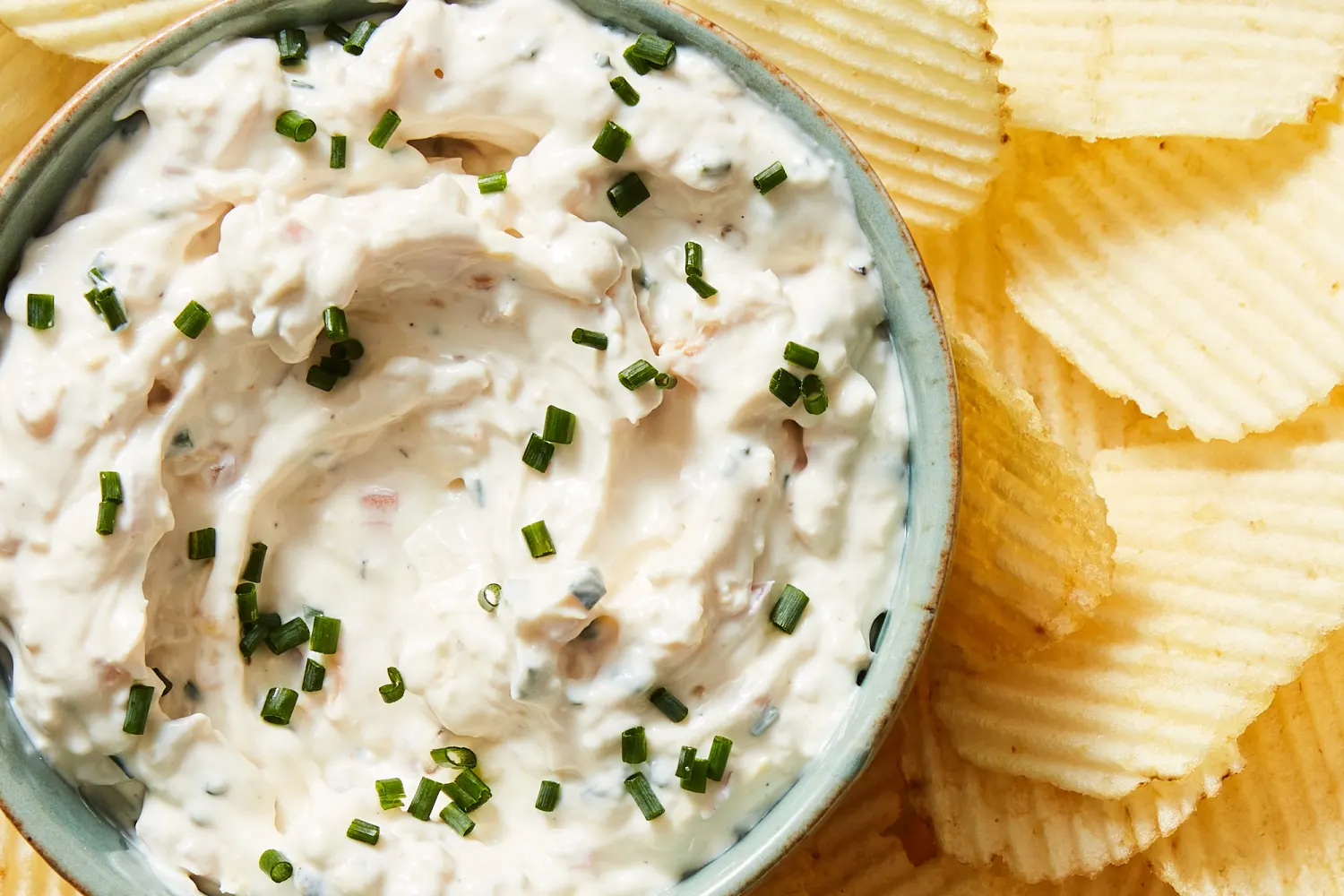 A bowl of clam dip on a bright kitchen counter surrounded by wavy potato chips and buttery crackers, ready for snacking