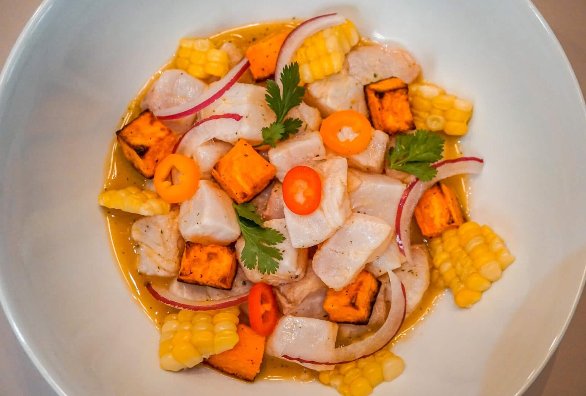 A bowl of classic Peruvian ceviche with white fish chunks, red onion slivers, cilantro, and a slice of lime, served with sweet potato and corn on a neutral stoneware plate on a bright kitchen table