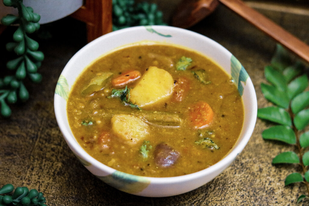 A bowl of classic South Indian sambar with visible vegetables and curry leaves, served beside fluffy white rice on a warm-toned kitchen table
