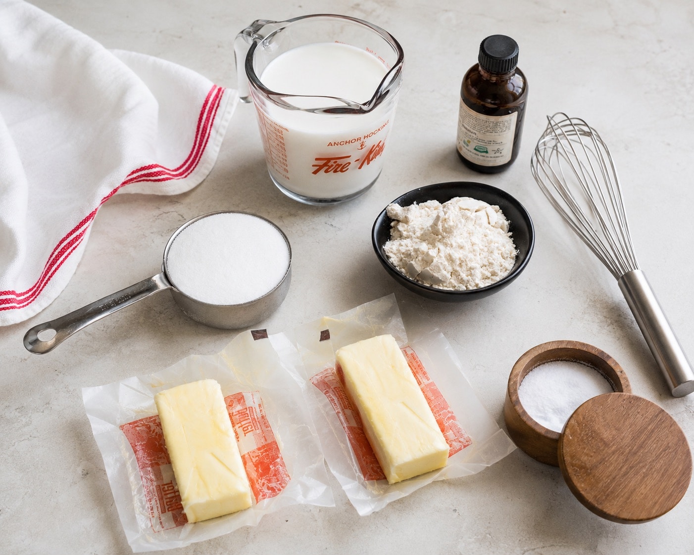 A bowl of cold heavy cream, a bowl of powdered sugar, vanilla extract, and a block of cream cheese arranged on a tidy kitchen counter