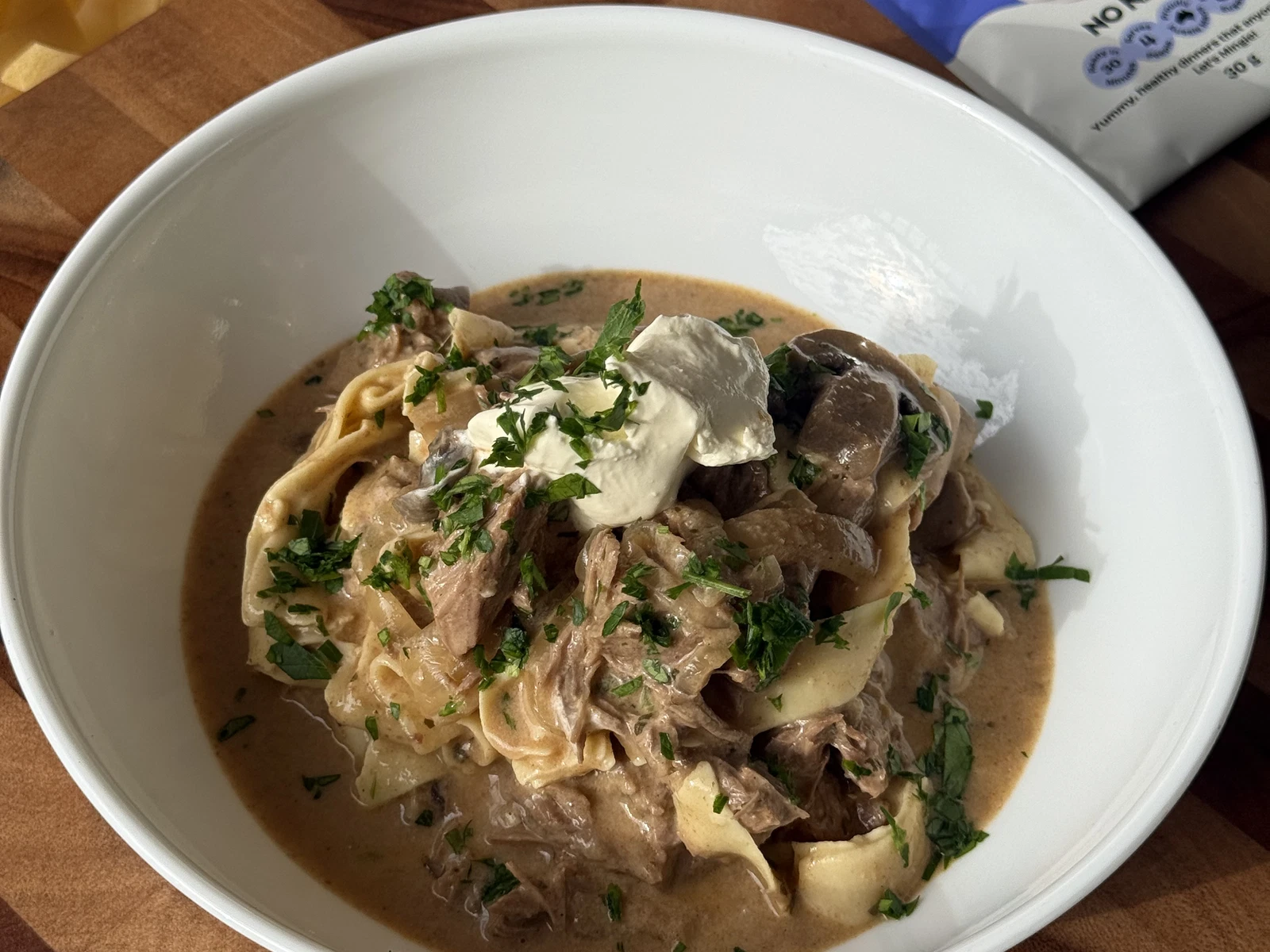 A bowl of creamy beef stroganoff with tender beef pieces and mushrooms served over egg noodles, garnished with parsley on a rustic table