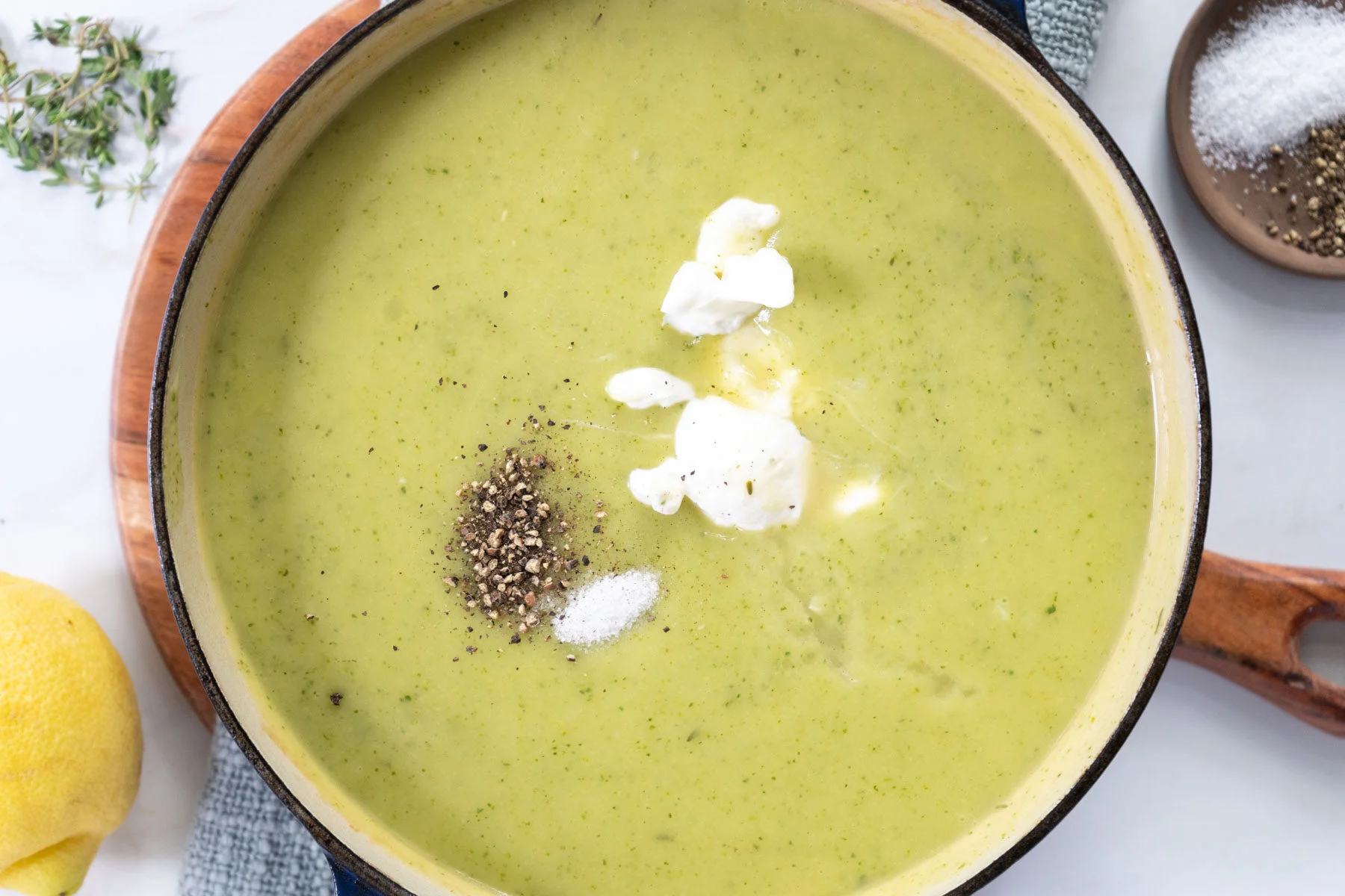 A bowl of creamy zucchini soup topped with olive oil, cracked black pepper, and chopped herbs on a light kitchen table with a spoon and napkin