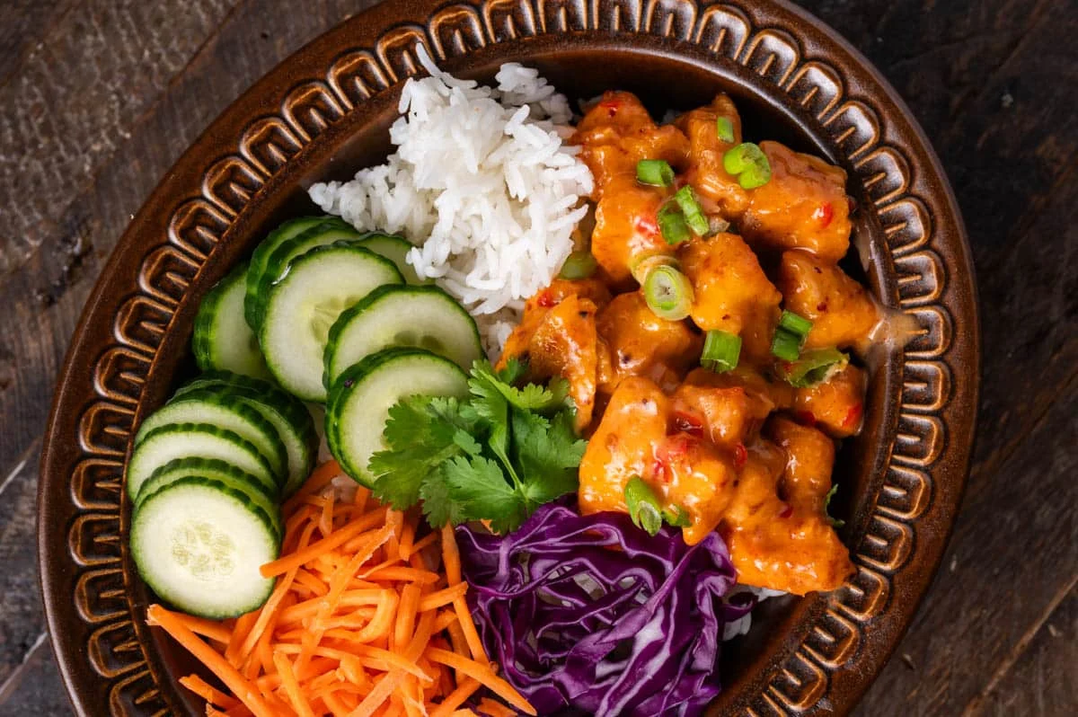 A bowl of crispy bang bang chicken bites coated in glossy creamy sauce, topped with sliced green onions and sesame seeds on a neutral kitchen table with soft natural light