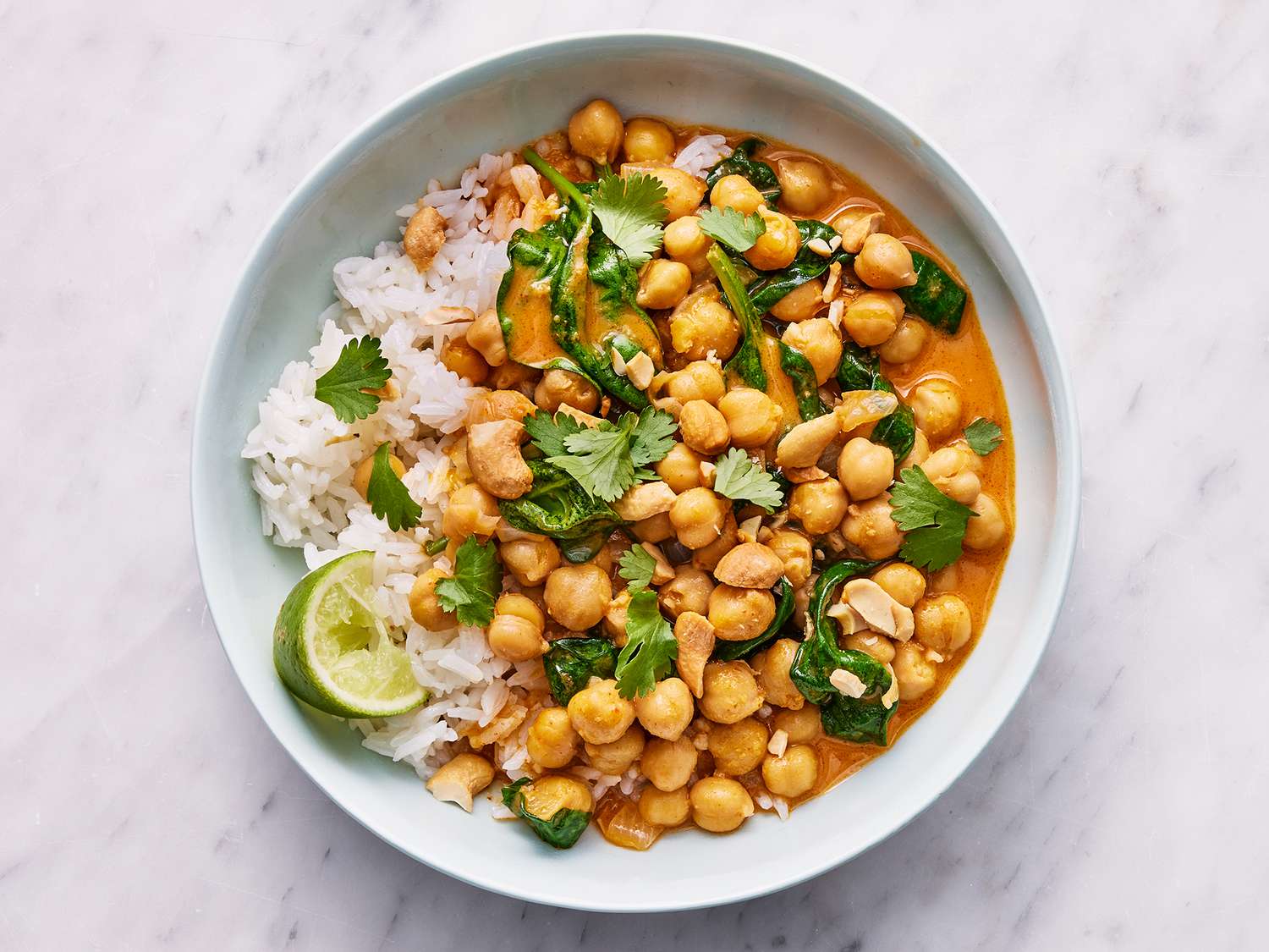A bowl of curry garbanzo beans in a rich orange coconut-tomato sauce, topped with cilantro and served with fluffy basmati rice on a warm-toned kitchen table