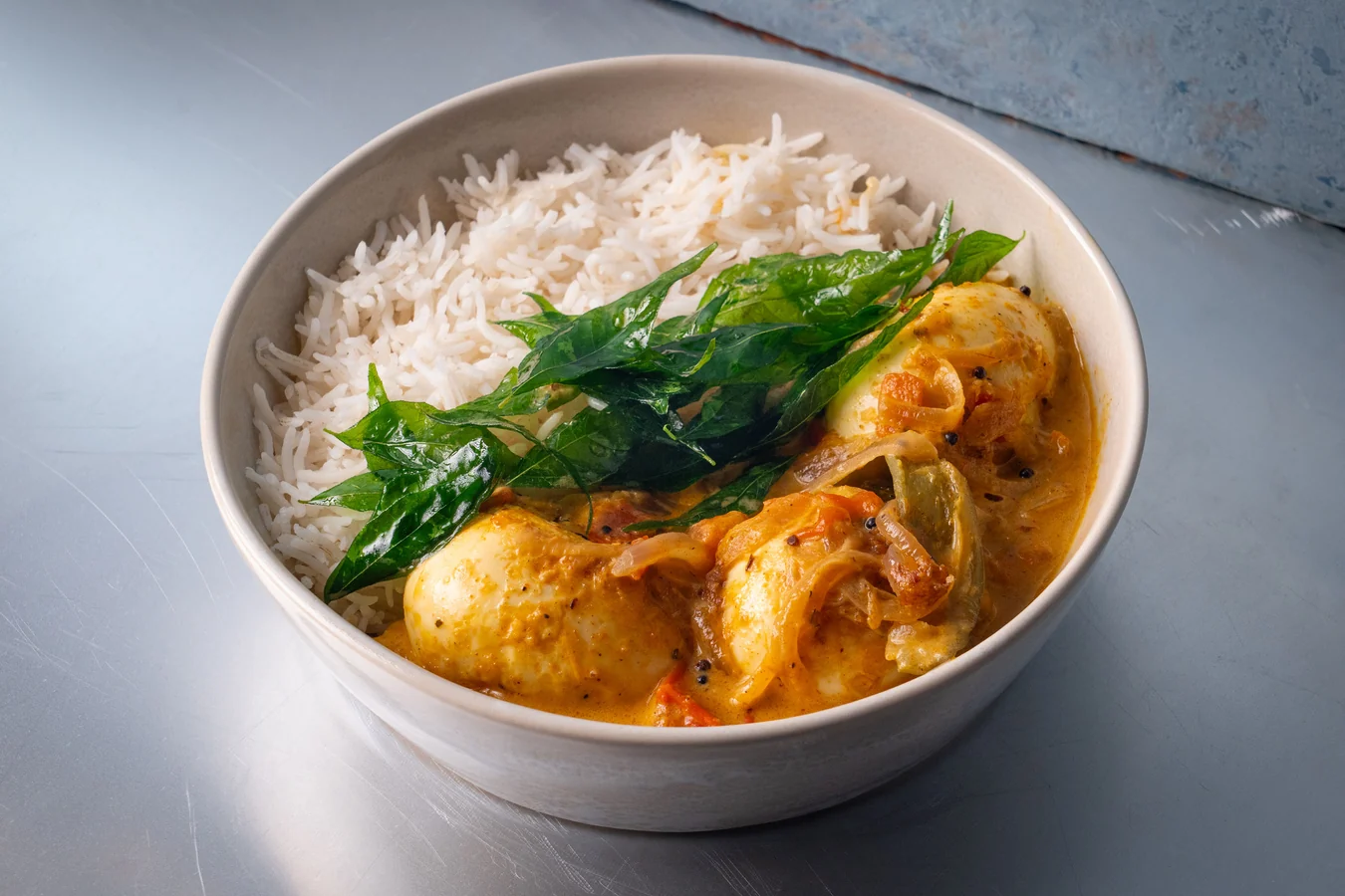 A bowl of egg curry with halved boiled eggs in a rich red-orange gravy, garnished with cilantro, served with basmati rice on a warm, softly lit kitchen table