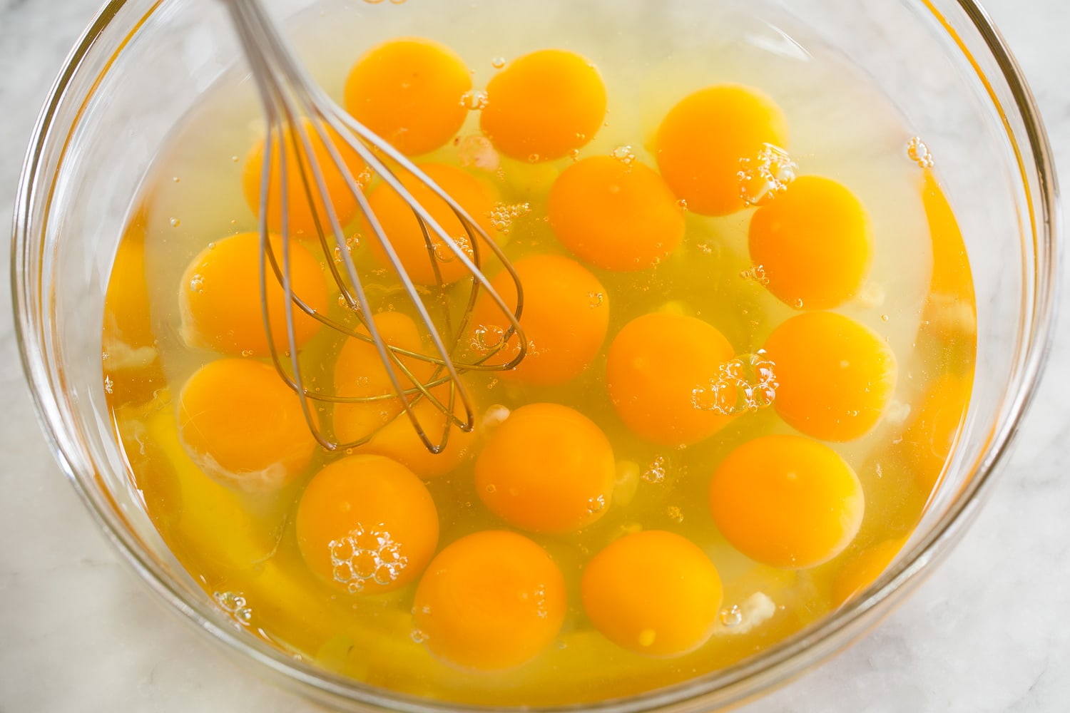 A bowl of eggs being whisked with milk, salt, pepper, and paprika on a kitchen counter with a whisk and measuring cup nearby