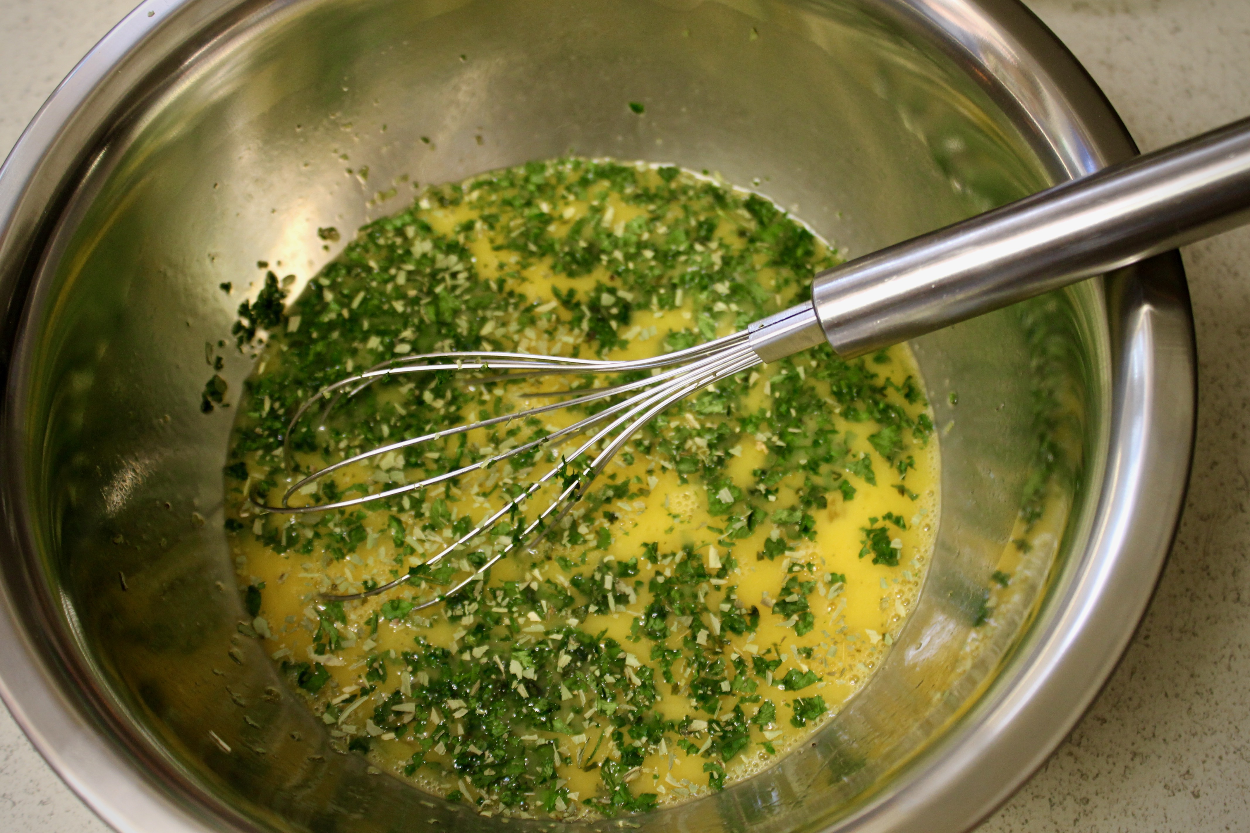 A bowl of eggs beside chopped fresh herbs, a stick of butter, and a lemon on a bright kitchen counter