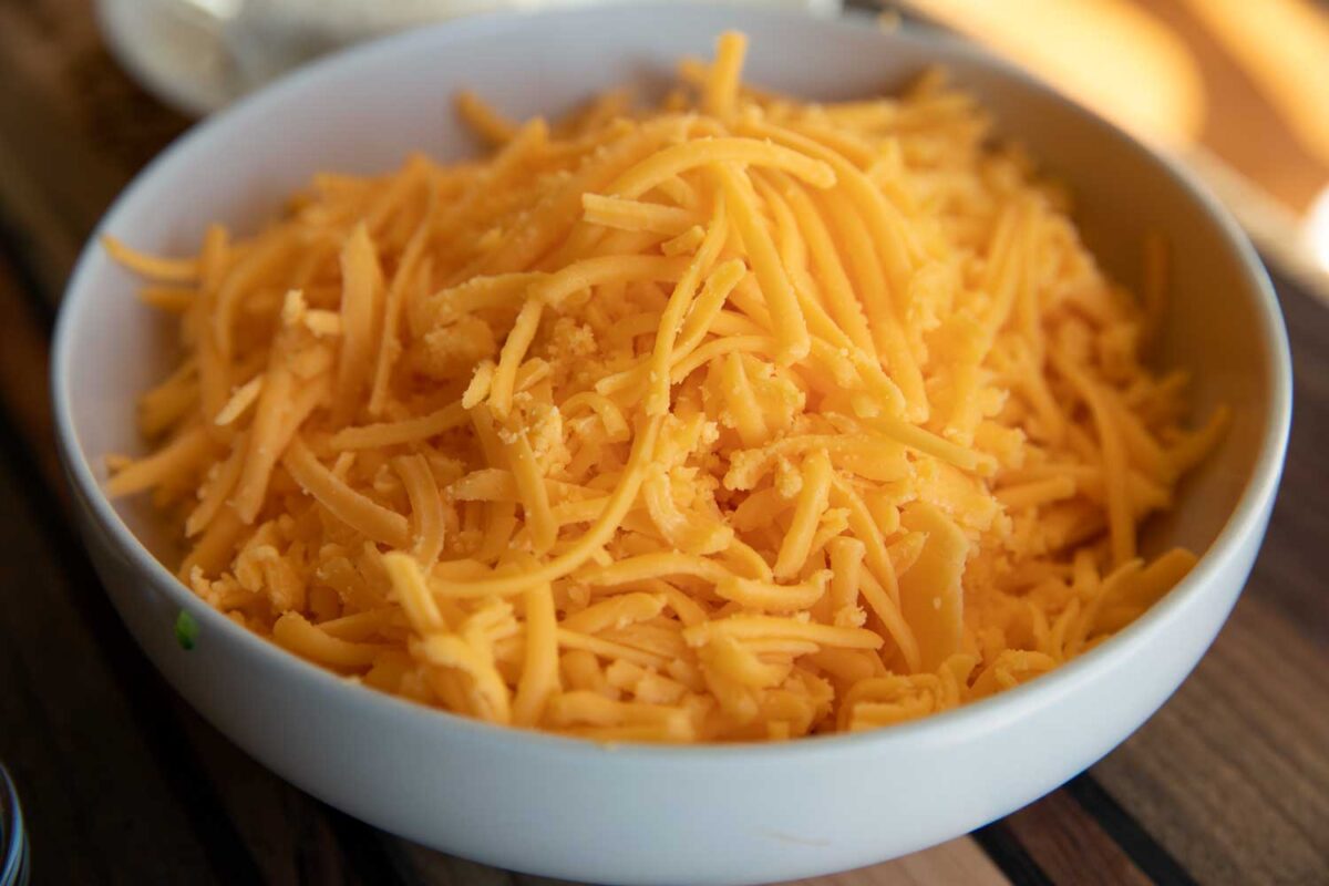 A bowl of freshly shredded sharp cheddar cheese on a kitchen counter next to a box grater, ready for biscuit dough