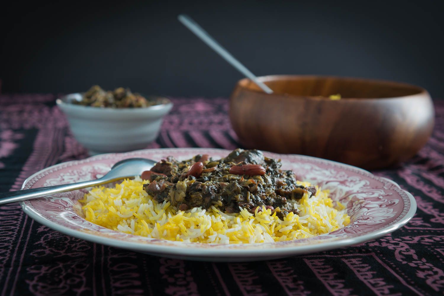 A bowl of ghormeh sabzi with dark green herb stew and tender beef chunks served beside fluffy basmati rice with a saffron-tinted top, on a warm kitchen table with soft natural light