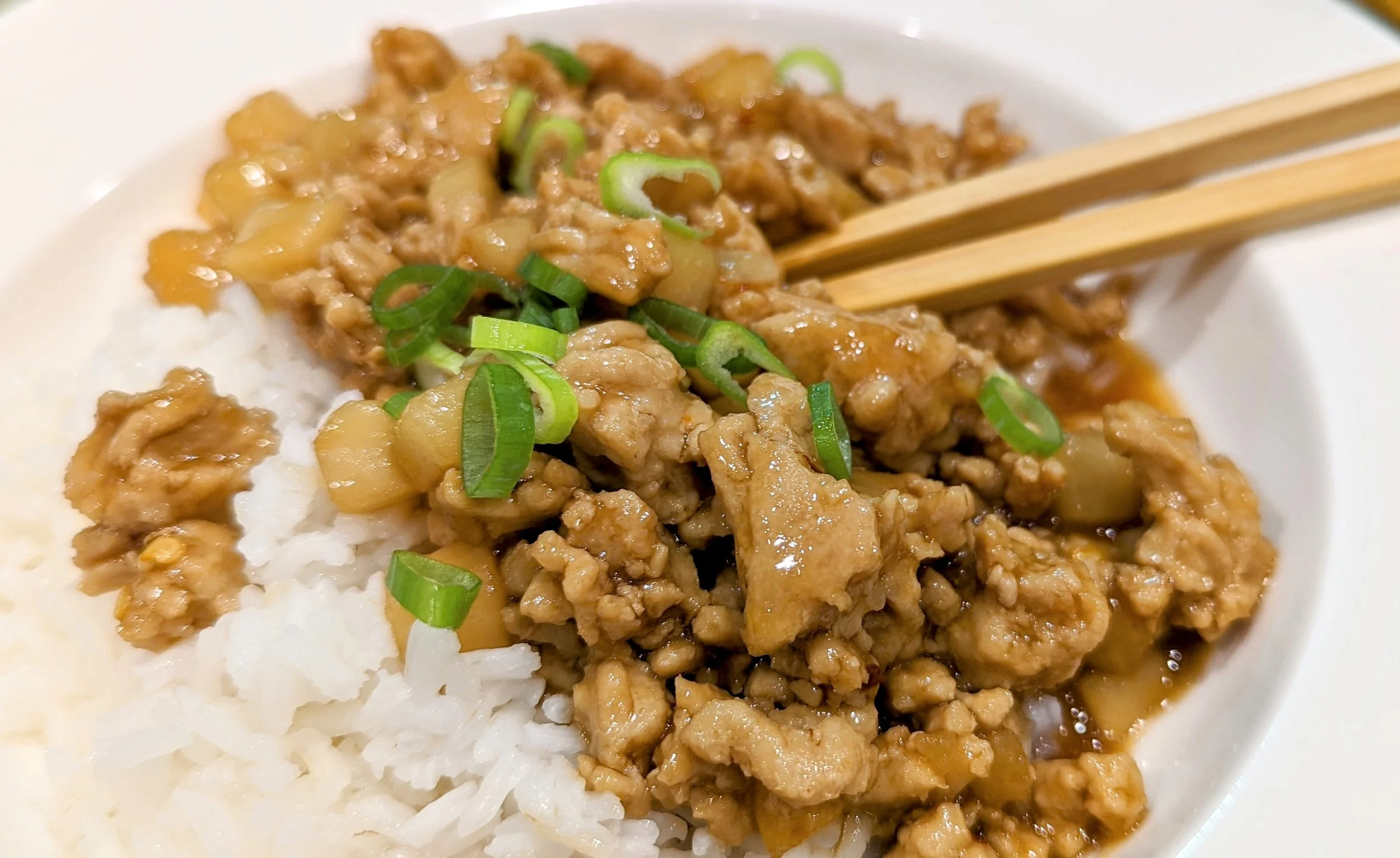 A bowl of glossy General Joe's chicken with sesame seeds and sliced scallions, served over steamed rice on a warm-toned kitchen table with chopsticks and a simple napkin