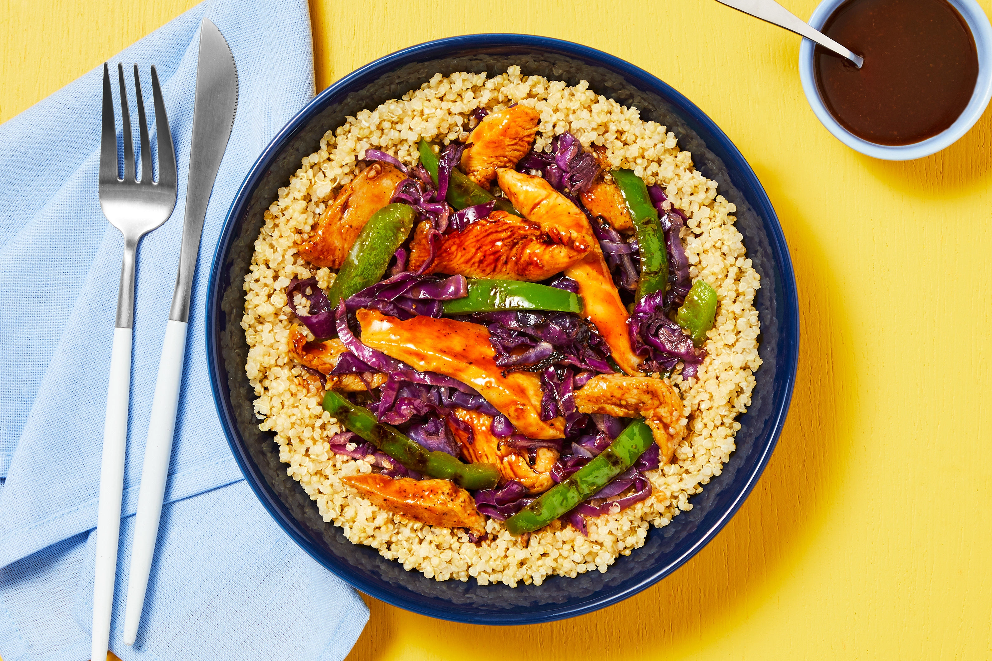 A bowl of glossy Szechuan chicken with red bell peppers and scallions, served over steamed white rice on a warm-toned kitchen table with chopsticks