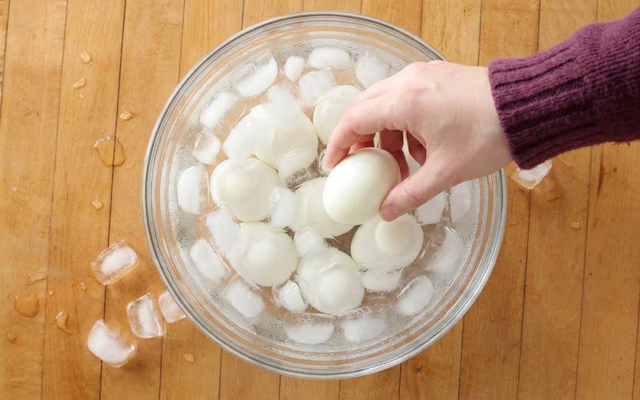 A bowl of hard-boiled eggs sitting in an ice bath on a kitchen counter, with condensation and scattered ice cubes