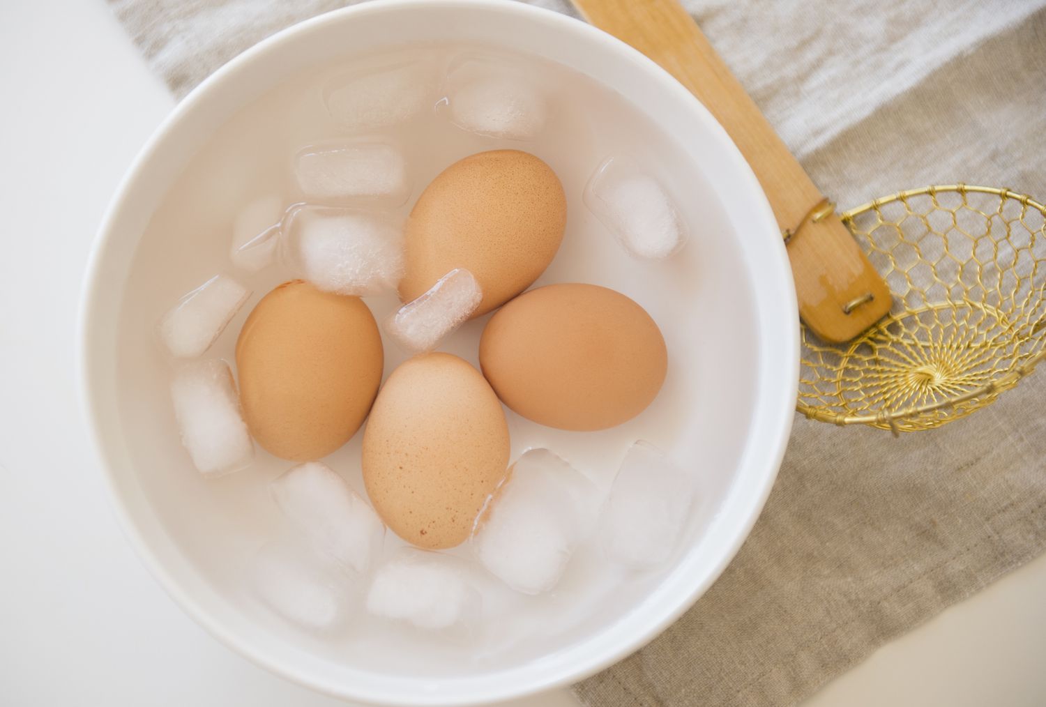 A bowl of ice water next to several eggs on a clean kitchen counter with a saucepan nearby