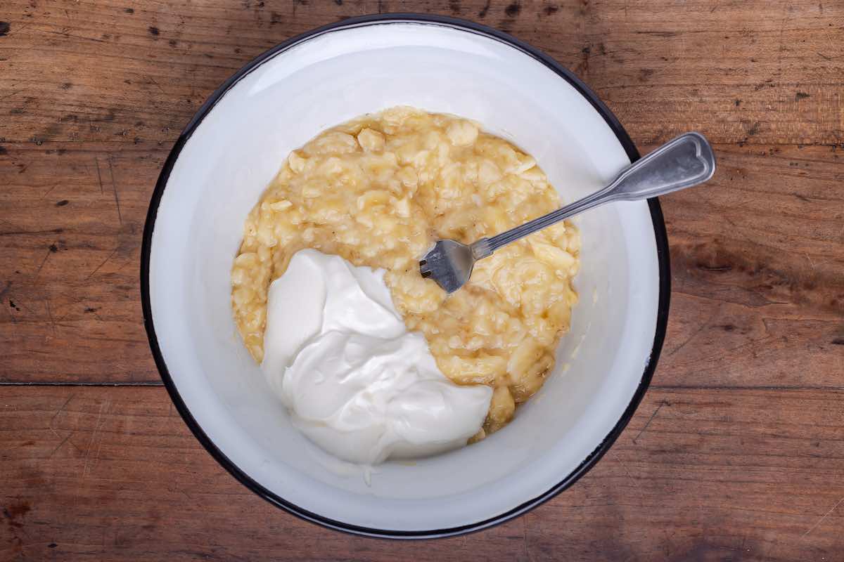 A bowl of mashed ripe bananas with a dollop of plain yogurt on top, ready to be mixed on a kitchen counter