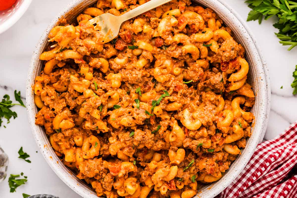 A bowl of old fashioned American goulash with elbow macaroni and ground beef in a rich tomato sauce, topped with a sprinkle of parsley on a simple dinner table
