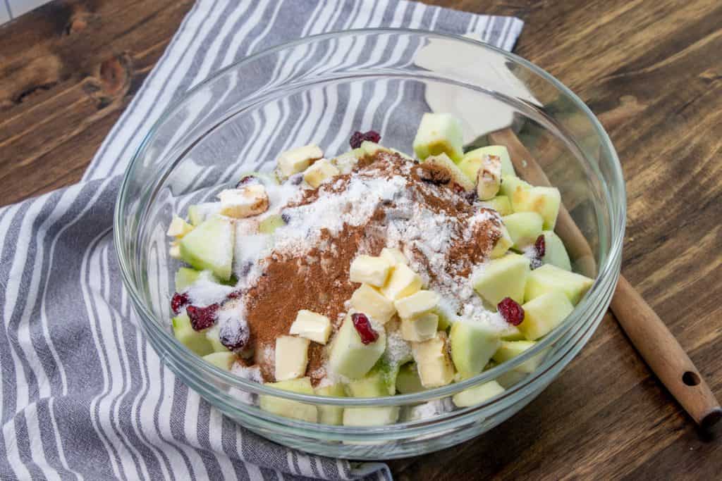A bowl of peeled diced apples on a kitchen counter with flour and cinnamon, with a spoon nearby