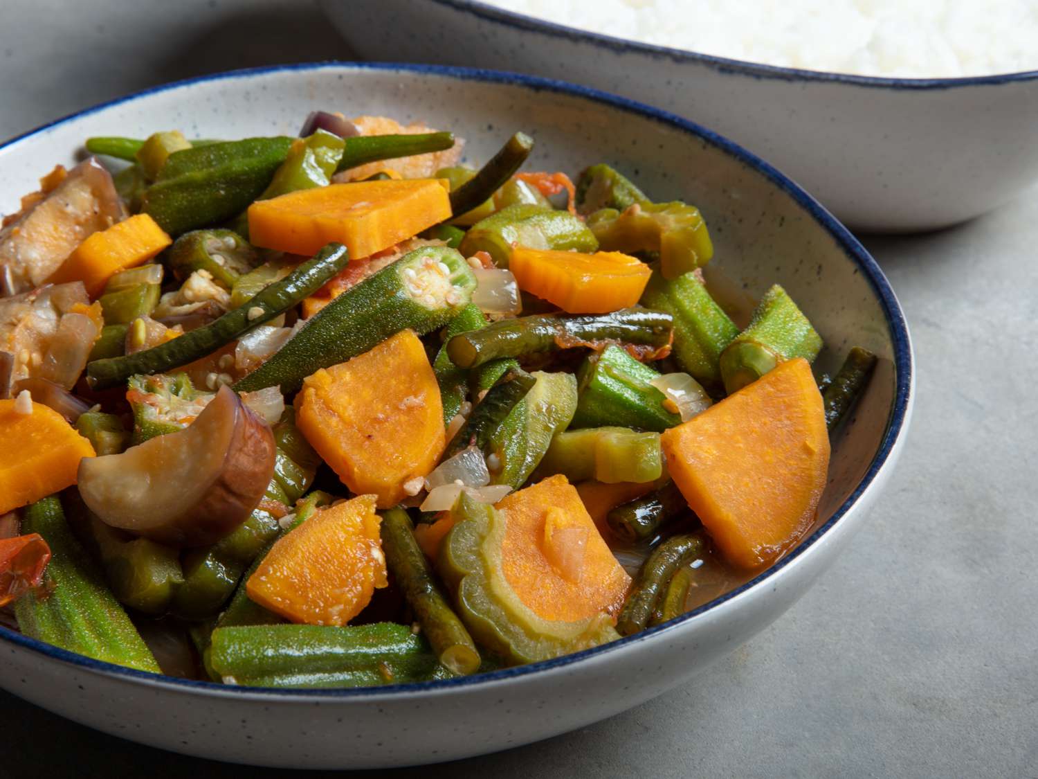 A bowl of pinakbet with squash, eggplant, okra, and green beans in a savory sauce, served with steamed rice on a warm kitchen table