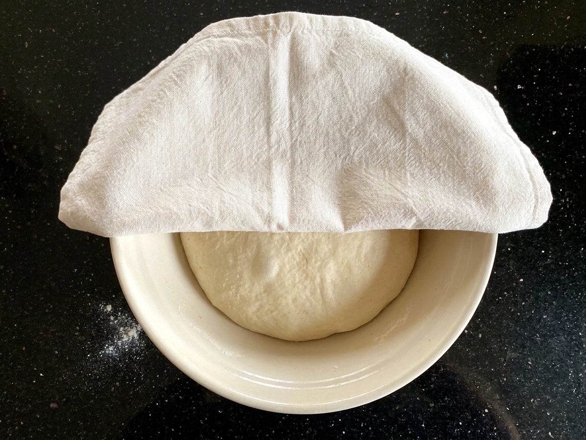 A bowl of pizza dough covered with a towel rising on a kitchen counter in warm light