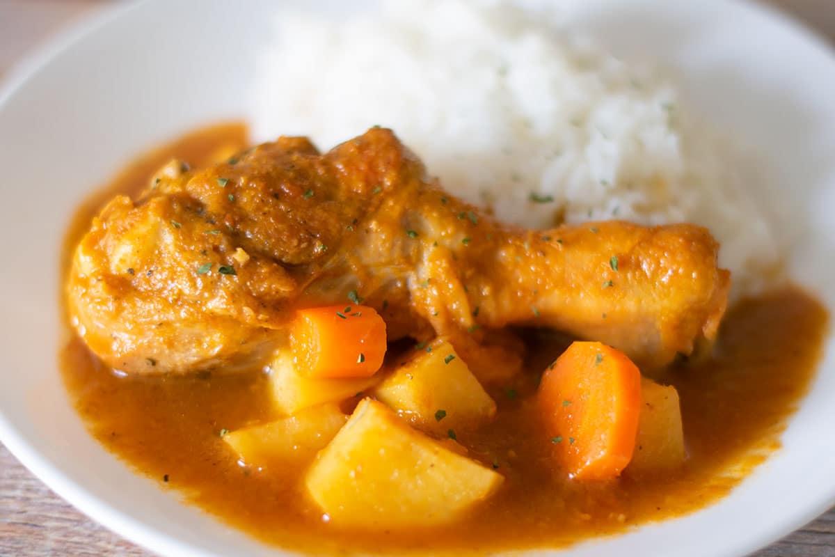 A bowl of pollo guisado with tender chicken and potatoes in a rich tomato gravy, served next to fluffy white rice on a simple wooden table