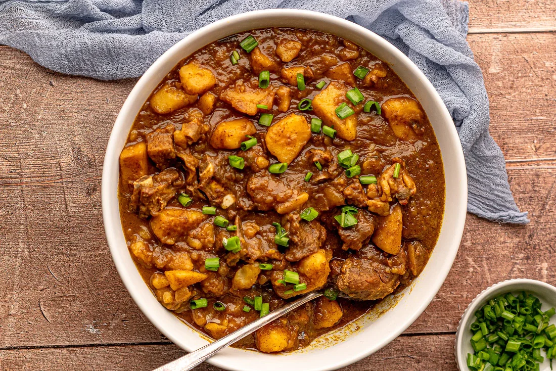 A bowl of rich goat curry with tender chunks of meat in a deep orange-brown coconut sauce, garnished with cilantro and lime wedges on a wooden table