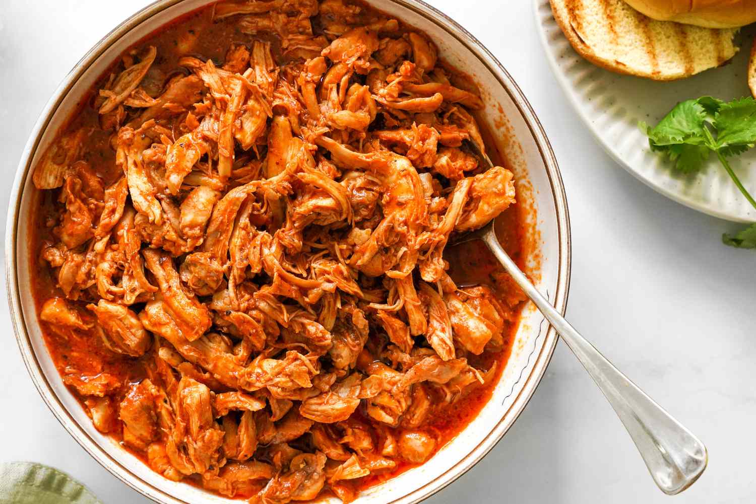 A bowl of shredded cooked chicken being tossed with BBQ sauce using a spoon on a kitchen counter