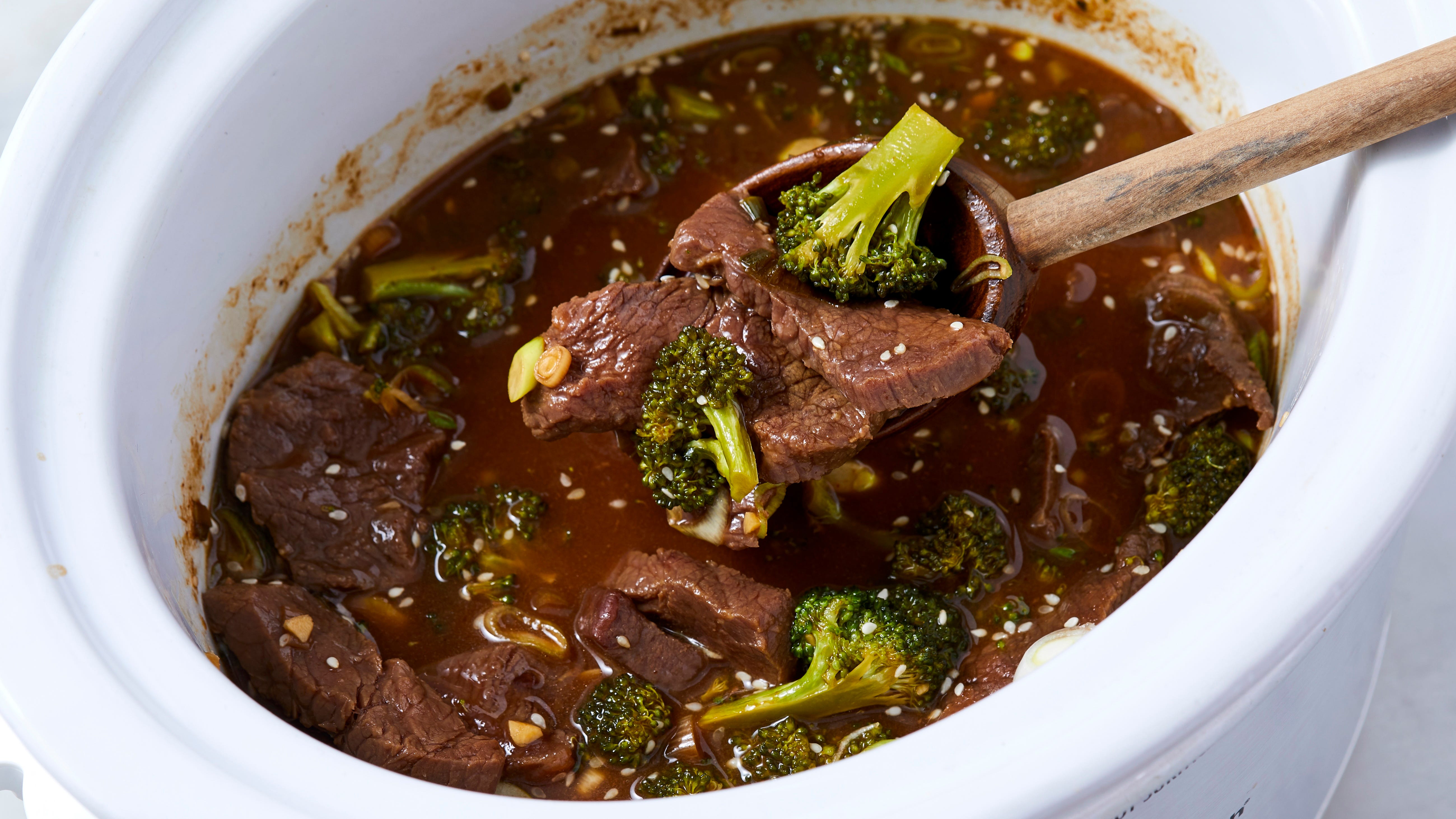 A bowl of slow cooker beef and broccoli in a glossy brown garlic sauce served over white rice, with sesame seeds and sliced green onions on top, on a warm wood table