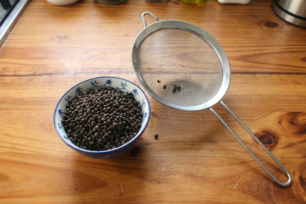 A bowl of soaked whole black lentils with water drained, sitting beside a fine mesh strainer on a kitchen counter