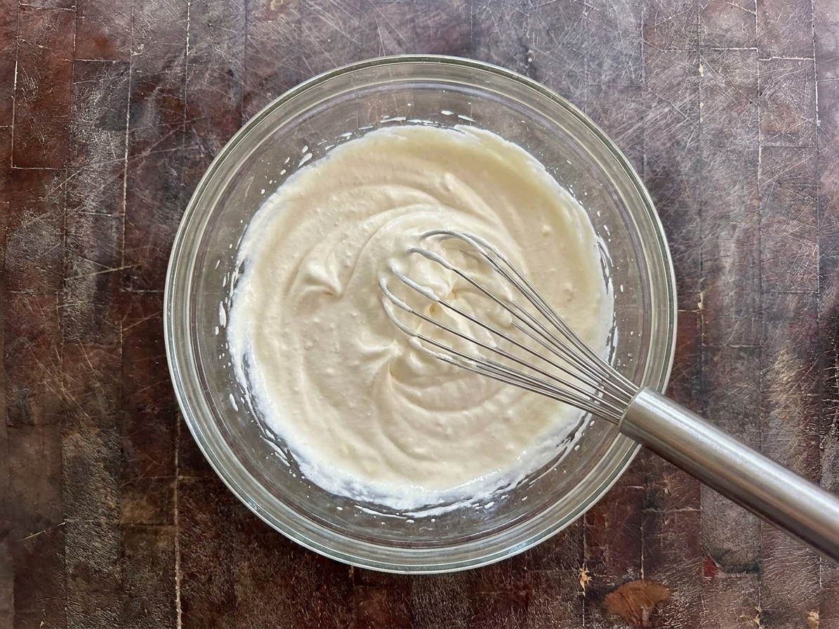 A bowl of softly whipped cream with a whisk resting inside on a bright kitchen counter