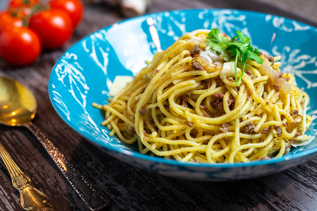 A bowl of spaghetti topped with rich minced beef tomato sauce, grated Parmesan, and fresh basil on a wooden table with a fork and napkin