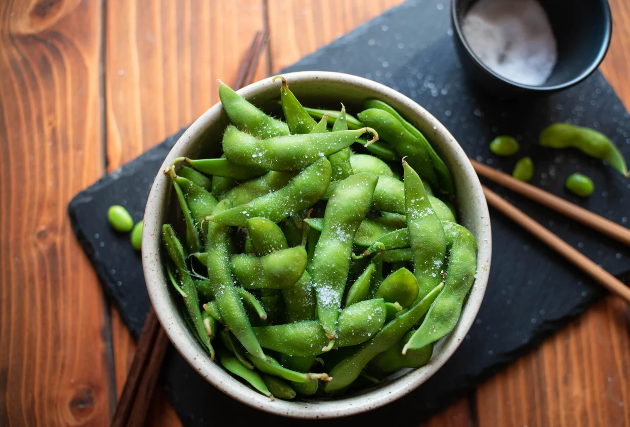 A bowl of steamed edamame pods sprinkled with flaky salt, served on a wooden table, photorealistic food photography