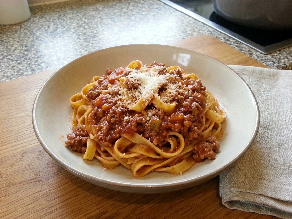A bowl of tagliatelle coated in traditional bolognese ragù with a dusting of Parmigiano-Reggiano on a wooden table with a linen napkin