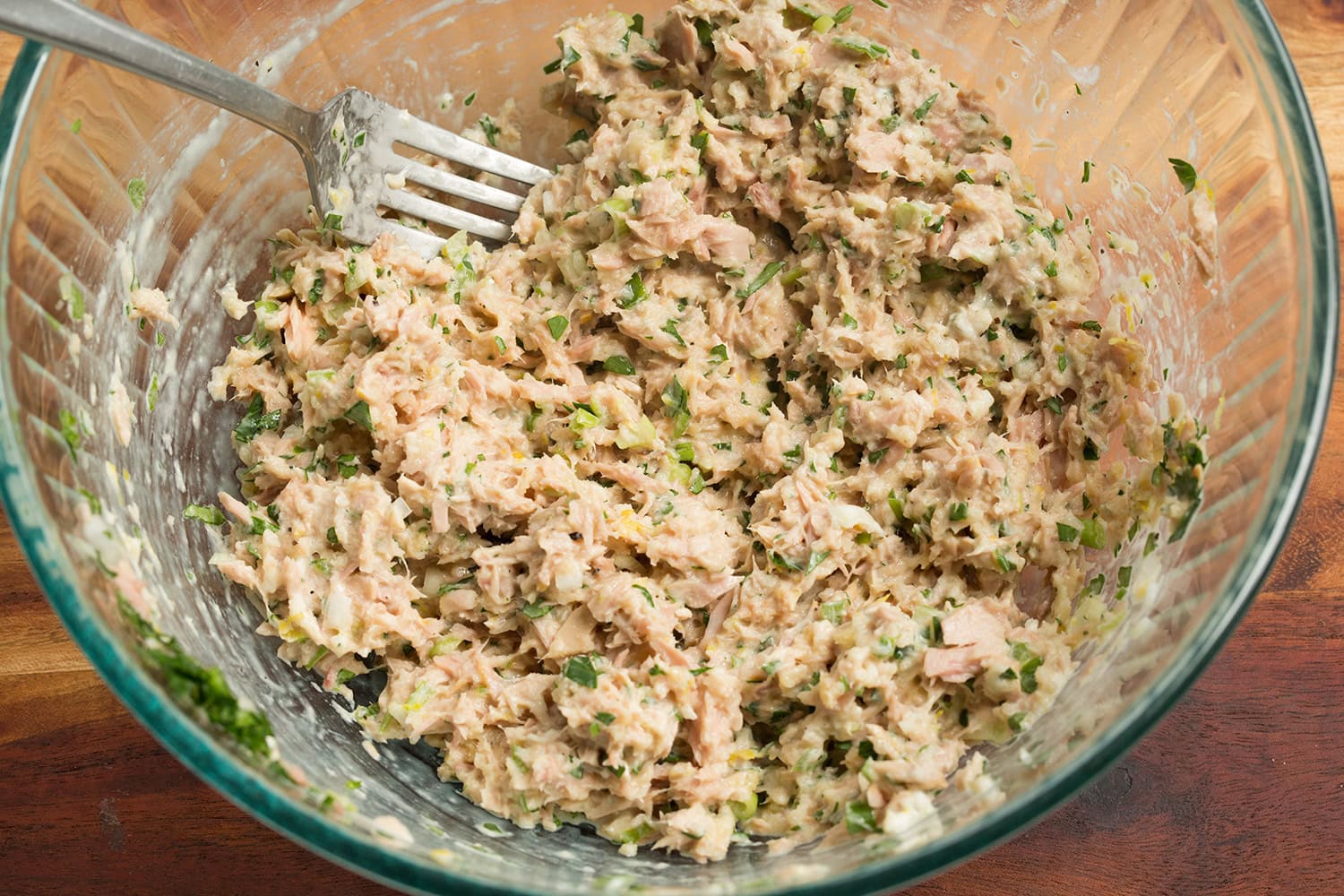 A bowl of tuna patty mixture being stirred with a fork on a kitchen counter, showing breadcrumbs, herbs, and diced onion
