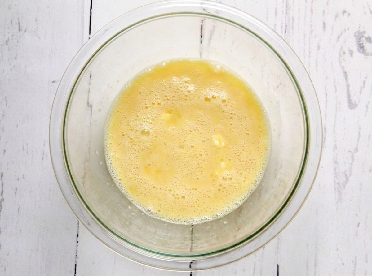 A bowl of whisked eggs, pale yellow and lightly foamy, on a wooden countertop