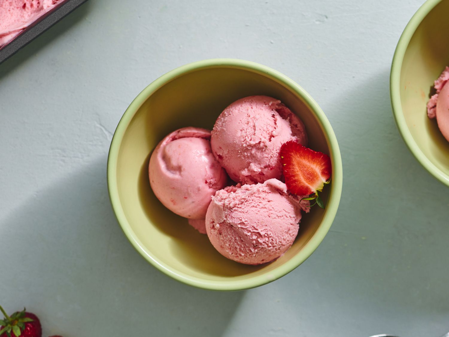 A bowl with two scoops of homemade strawberry ice cream with visible strawberry pieces, set on a light kitchen table with a spoon nearby, natural window light