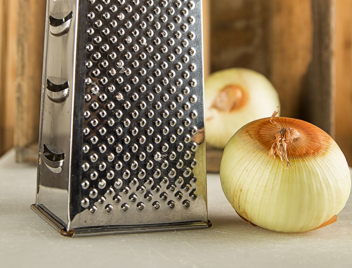 A box grater with freshly grated onion on a cutting board beside a mixing bowl, in a bright home kitchen