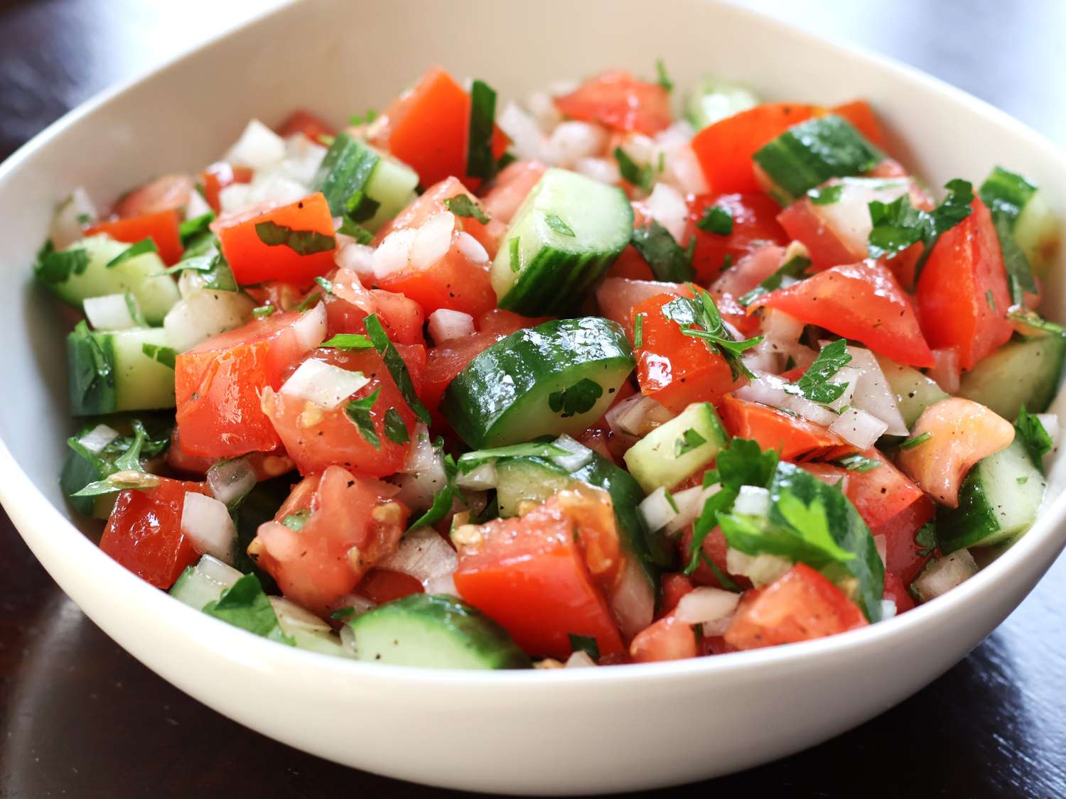 A bright bowl of cucumber and tomato salad with red onion and fresh herbs on a wooden kitchen table in natural window light