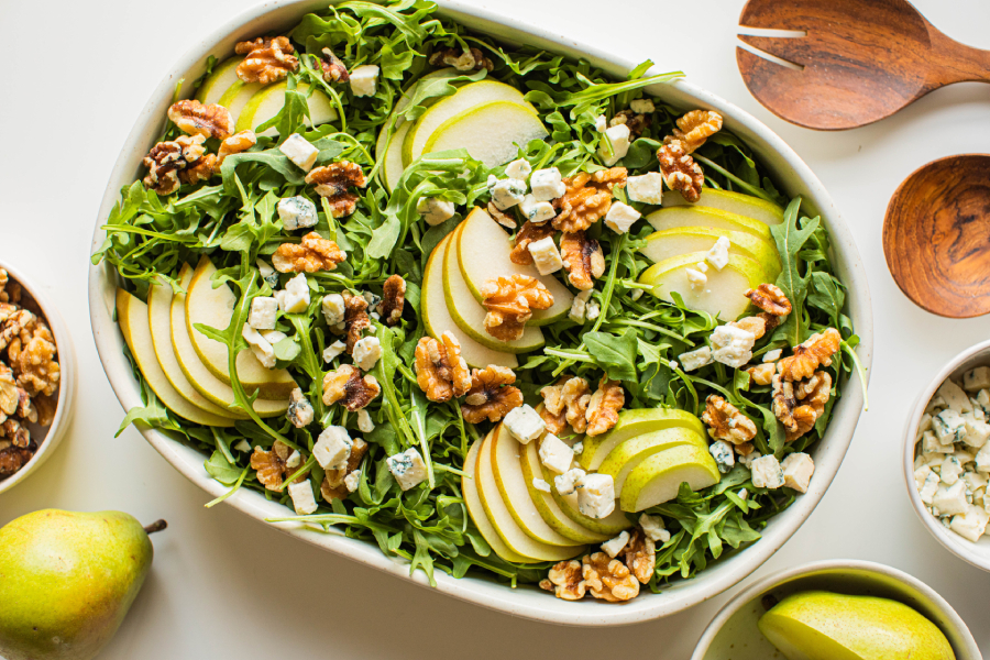 A bright plated pear salad on a white serving platter with arugula, sliced ripe pears, toasted pecans, crumbled blue cheese, and a light vinaigrette sheen on a wooden table in natural window light