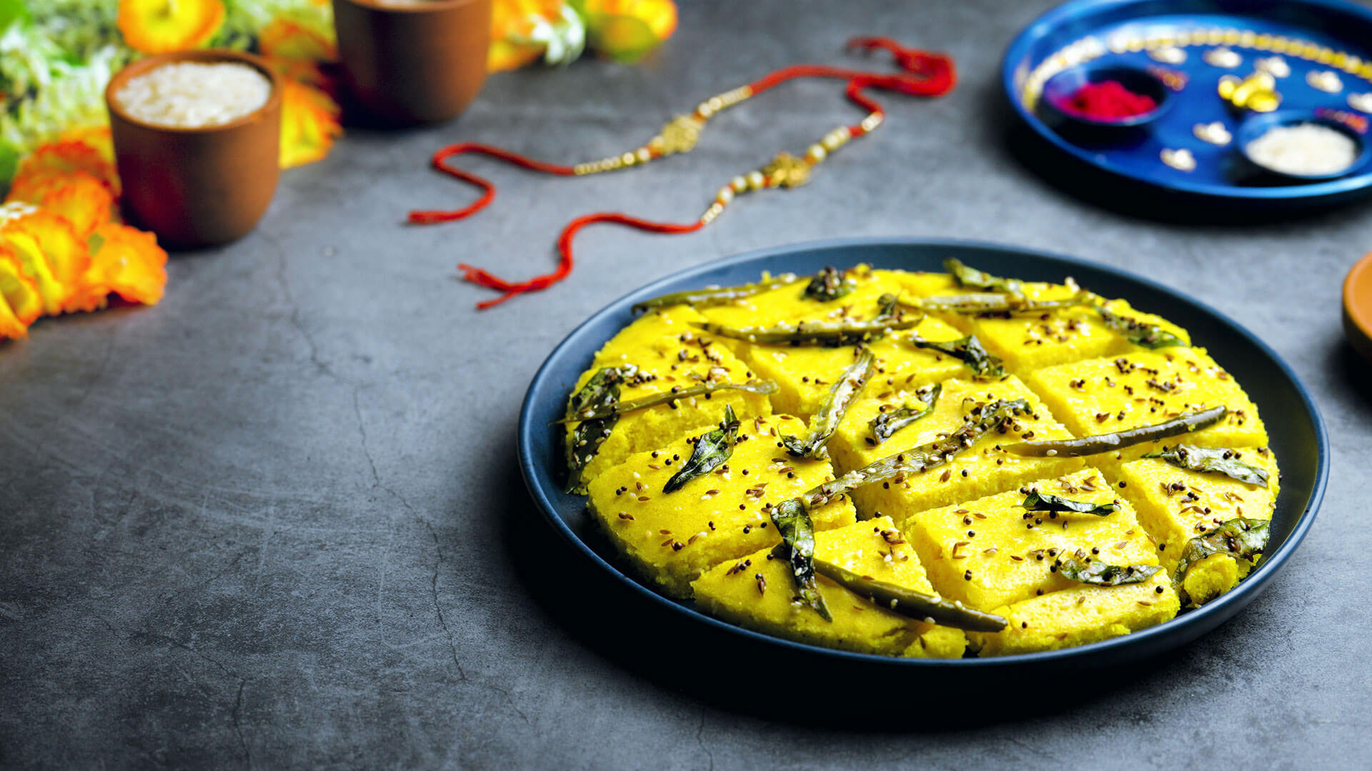 A bright yellow tray of sliced khaman dhokla topped with mustard seeds, curry leaves, and green chiles on a light kitchen counter