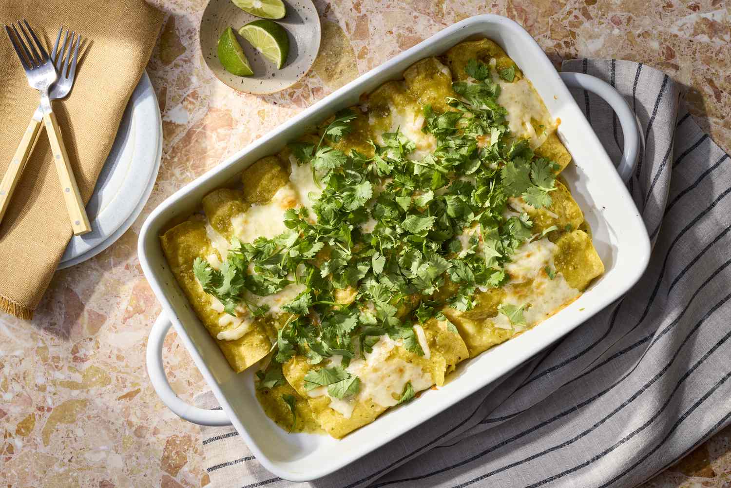 A bubbling baking dish of green chicken enchiladas covered in melted cheese, salsa verde, and scattered cilantro on a light stone kitchen counter