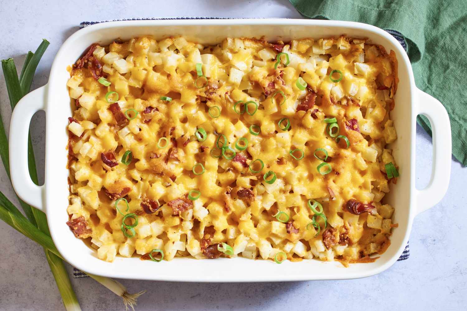 A bubbling casserole dish of Mississippi mud potatoes with melted cheddar on top, crispy bacon bits, and sliced green onions on a rustic wooden table