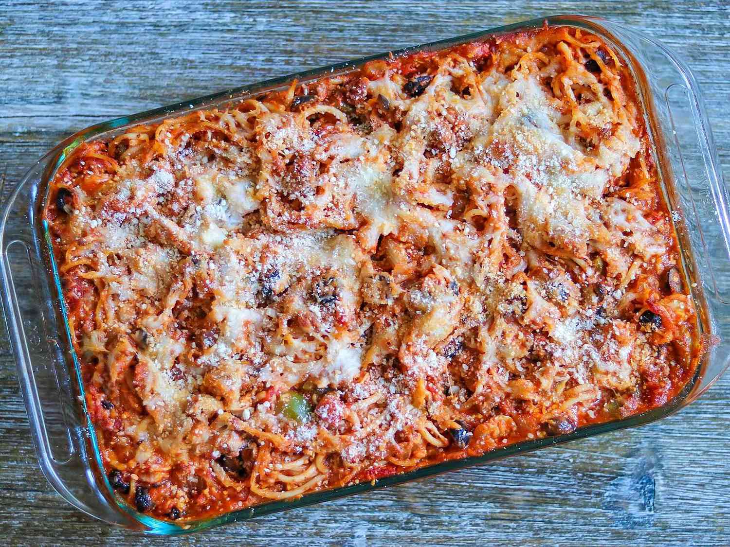 A bubbling casserole dish of baked spaghetti topped with melted mozzarella and golden brown cheese spots, sitting on a light stone countertop with a wooden spoon nearby