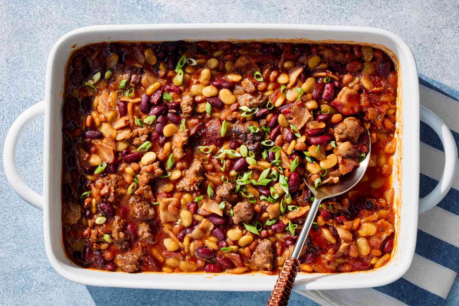 A bubbling casserole dish of calico beans with visible bacon pieces and mixed beans in a glossy sweet-tangy sauce on a warm-toned kitchen counter