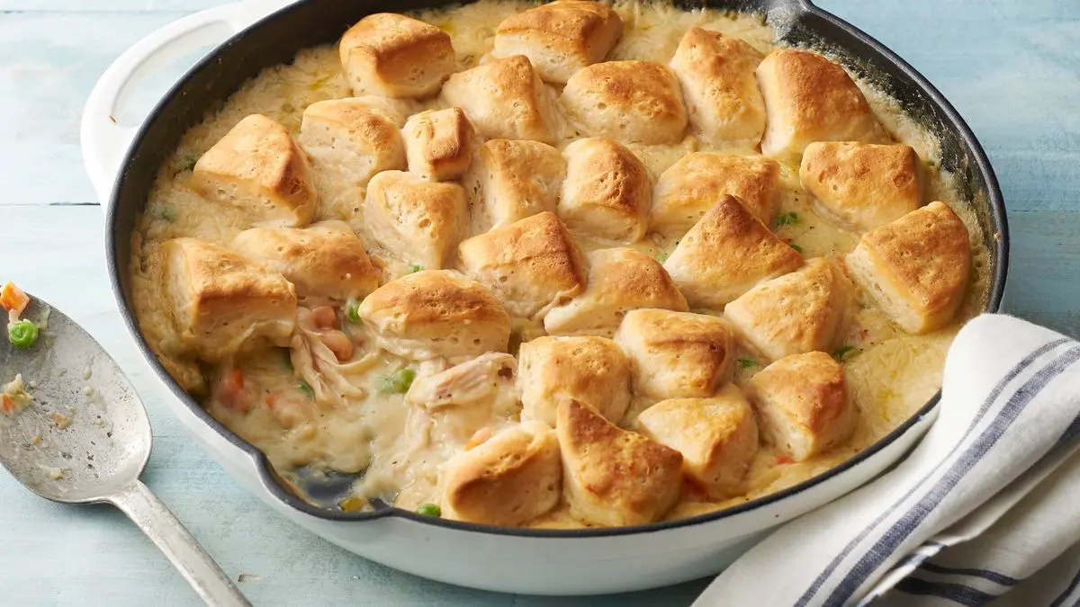 A bubbling chicken pot pie casserole in a white baking dish topped with golden Pillsbury biscuits, set on a warm kitchen table with simple plates