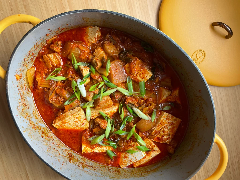 A bubbling pot of kimchi stew with pork and tofu on a stovetop, steam rising, with a bowl of white rice and small side dishes on a warm kitchen table