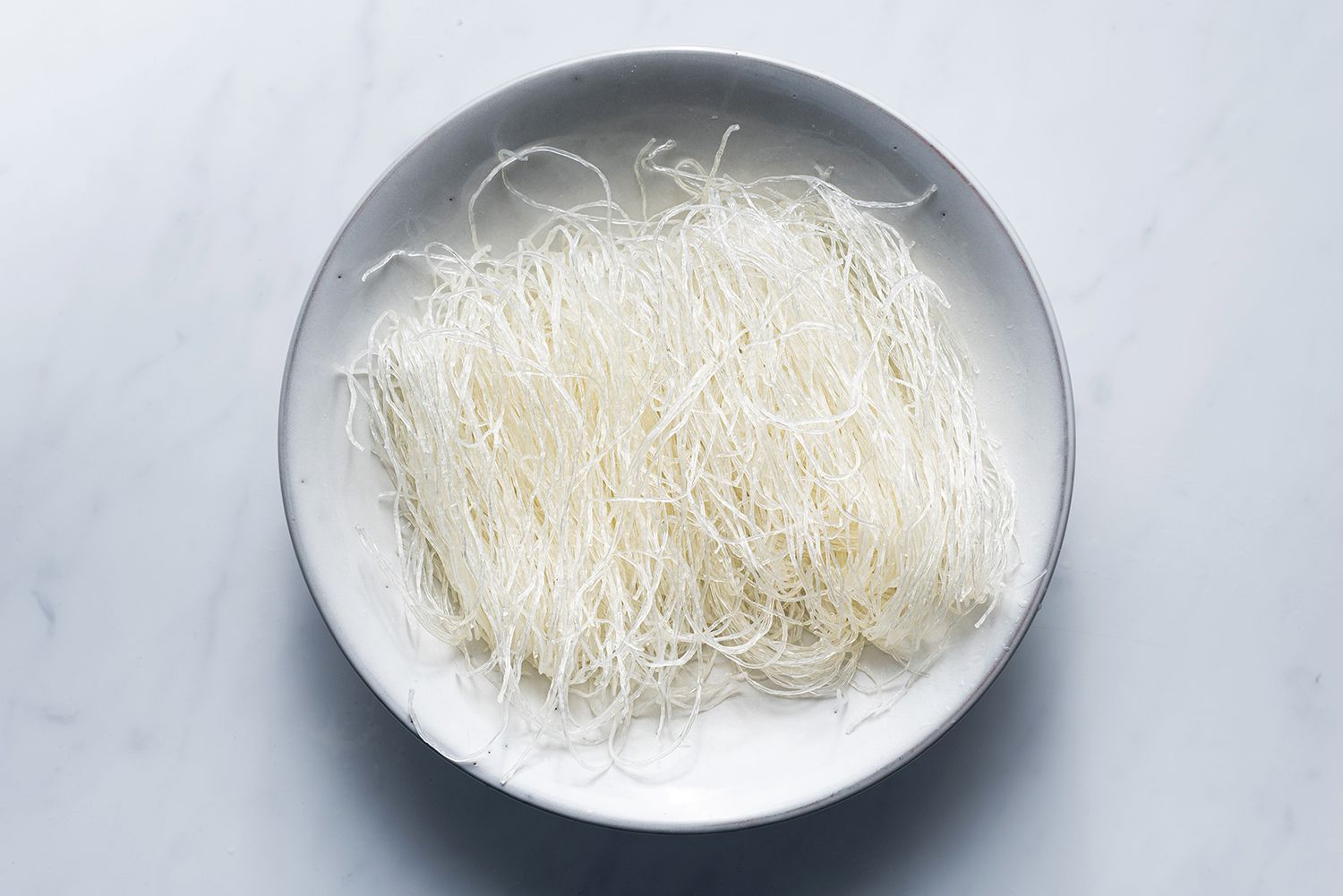 A bundle of dried glass noodles on a wooden cutting board next to small bowls of soy sauce, sesame oil, garlic, and sliced vegetables