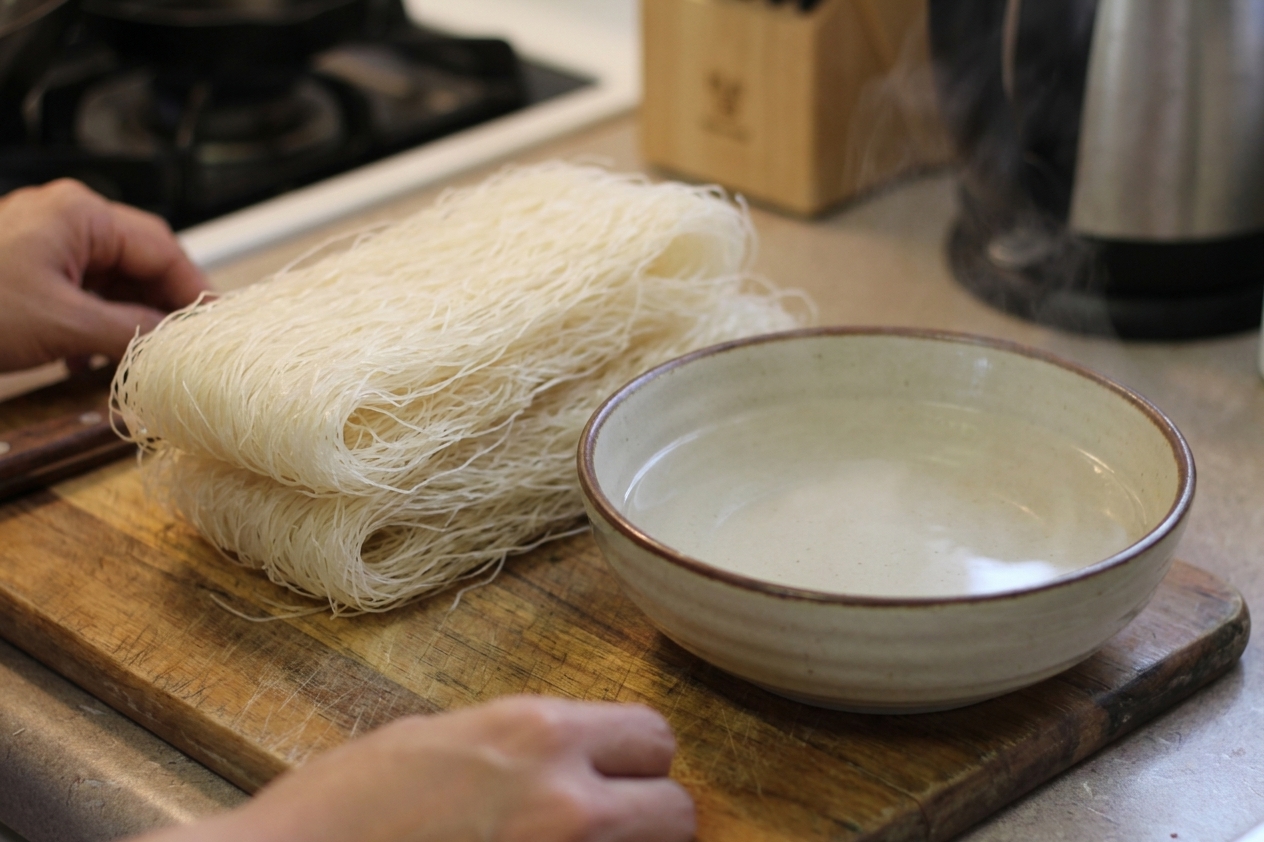 A bundle of dry bihon rice vermicelli noodles on a wooden cutting board next to a bowl of warm water