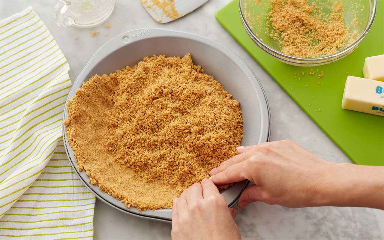 A buttery graham cracker crumb mixture being pressed firmly into the bottom of a springform pan on a kitchen counter