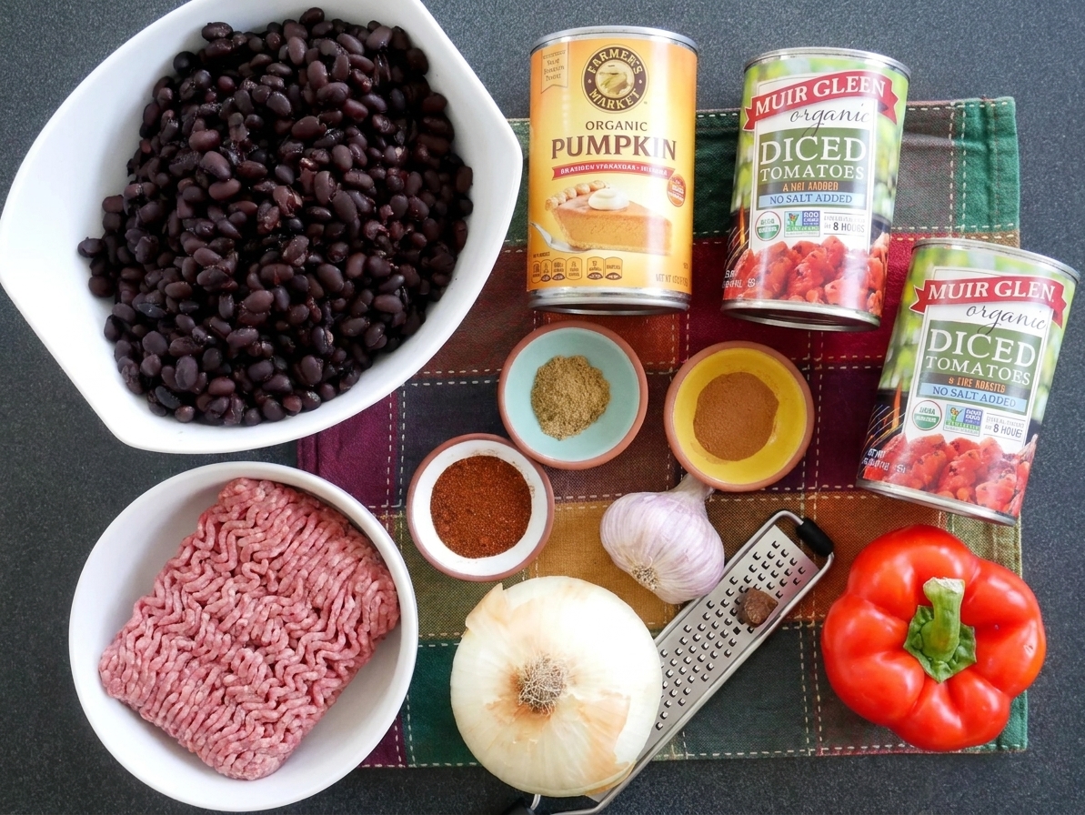 A can of pumpkin puree alongside beans, diced tomatoes, onions, garlic, ground meat, and spices set out on a clean kitchen counter before cooking