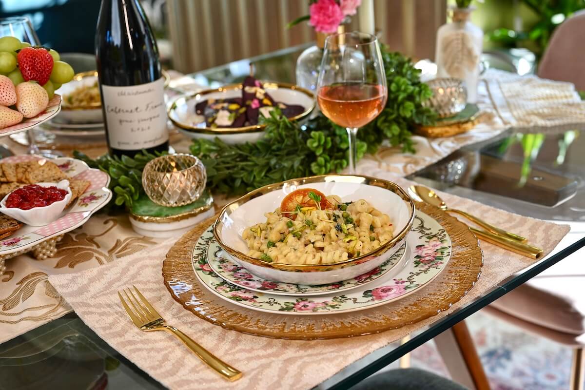 A candlelit dinner table set with a shallow bowl of lemon herb pasta topped with crispy breadcrumbs, a small vase of greenery, linen napkins, and warm evening lighting